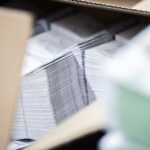 Mehrere sauber gefalzte Beipackzettel sind dicht an dicht in einem offenen Karton gestapelt. Die gleichmäßig ausgerichteten Papierkanten bilden eine klare Linienstruktur, während schwarze Heftklammern entlang des Rückens sichtbar sind. Der Blick erfolgt durch die Kartonöffnung, wodurch Vorder- und Hintergrund leicht unscharf erscheinen. Das Bild zeigt fertig produzierte Druckerzeugnisse, die für Lagerung oder Versand vorbereitet sind.