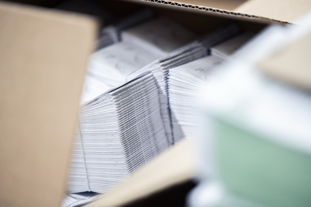 Mehrere sauber gefalzte Beipackzettel sind dicht an dicht in einem offenen Karton gestapelt. Die gleichmäßig ausgerichteten Papierkanten bilden eine klare Linienstruktur, während schwarze Heftklammern entlang des Rückens sichtbar sind. Der Blick erfolgt durch die Kartonöffnung, wodurch Vorder- und Hintergrund leicht unscharf erscheinen. Das Bild zeigt fertig produzierte Druckerzeugnisse, die für Lagerung oder Versand vorbereitet sind.