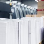 Große Papierstapel mit sauber geschnittenen Kanten sind auf Paletten in der Druckerei Esslinger Druck in Villingen-Schwenningen platziert. Die Druckbögen sind exakt übereinandergelegt und bereit für die Weiterverarbeitung oder Verpackung. Hinter den Stapeln stehen mehrere Druckmaschinen in Reihe, was auf eine Serienfertigung hindeutet. Die Szene zeigt professionelle Papierverarbeitung im industriellen Umfeld.