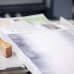 Ein dicker Stapel frisch gedruckter Bedienungsanleitungen liegt exakt ausgerichtet auf dem Arbeitstisch einer Schneidemaschine. Die feinen Papierkanten bilden eine gleichmäßige Linienstruktur, während im Hintergrund unscharf weitere Druckbögen zu erkennen sind. Neben dem Stapel liegt ein Holzklotz, der zur Fixierung beim Zuschnitt verwendet wird. Das Bild zeigt einen präzisen Arbeitsschritt in der Druckerei Esslinger Druck in Villingen-Schwenningen.