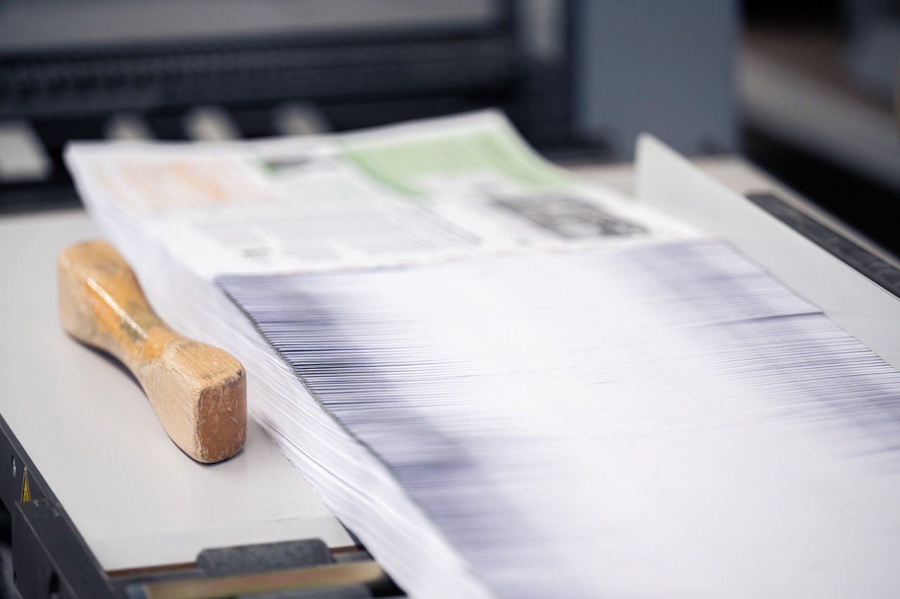 Ein dicker Stapel frisch gedruckter Bedienungsanleitungen liegt exakt ausgerichtet auf dem Arbeitstisch einer Schneidemaschine. Die feinen Papierkanten bilden eine gleichmäßige Linienstruktur, während im Hintergrund unscharf weitere Druckbögen zu erkennen sind. Neben dem Stapel liegt ein Holzklotz, der zur Fixierung beim Zuschnitt verwendet wird. Das Bild zeigt einen präzisen Arbeitsschritt in der Druckerei Esslinger Druck in Villingen-Schwenningen.