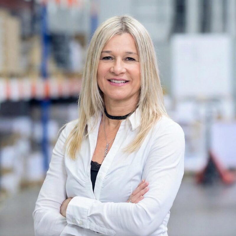 Eine blonde Frau mit einem schwarzen Choker und einer silbernen Halskette lächelt in die Kamera. Sie trägt eine weiße Bluse über einem schwarzen Oberteil und hat die Arme verschränkt. Im Hintergrund sind unscharfe Lagerregale und Paletten zu sehen.