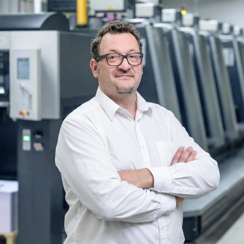 Ein lächelnder Mann mit Brille und kurzem Bart steht mit verschränkten Armen. Im Hintergrund sind grosse, graue Offsetdruckmaschinen und ein Stapel Papier auf einer Palette in einer Druckerei zu sehen.