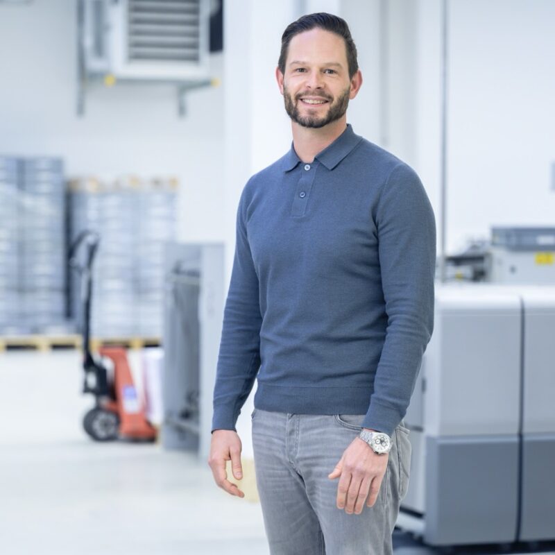 Ein lächelnder Mitarbeiter der Druckerei Esslinger Druck in Villingen-Schwenningen steht vor großen industriellen Druckmaschinen. Er trägt ein dunkelblaues Langarmshirt und graue Jeans und blickt selbstbewusst in die Kamera. Im Hintergrund sind Produktionsanlagen, Paletten und technische Geräte in einer hellen Halle zu sehen. Das Bild vermittelt Professionalität und Kompetenz im Bereich Druckproduktion.
