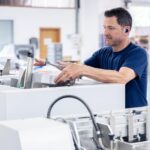 Ein Mann mit blauem T-Shirt positioniert einen dicken Stapel bedruckter Broschüren in einer automatisierten Papierverarbeitungsmaschine. Die Druckerzeugnisse sind exakt gestapelt und werden zwischen verstellbaren Anschlägen fixiert. Im Hintergrund sind weitere industrielle Geräte einer Druckerei sichtbar, die auf Serienproduktion hindeuten. Die Szene dokumentiert präzise Papierverarbeitung in der Druckerei Esslinger Druck in Villingen-Schwenningen.