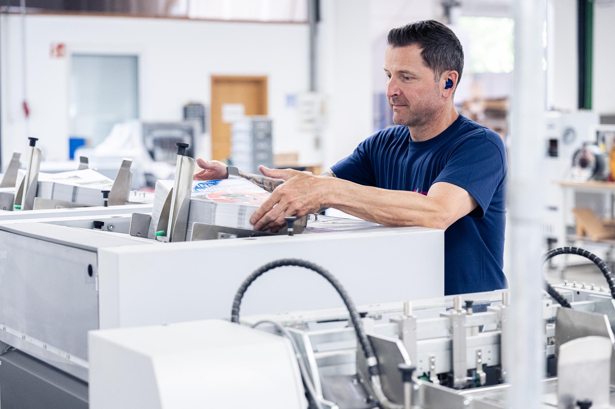 Ein Mann mit blauem T-Shirt positioniert einen dicken Stapel bedruckter Broschüren in einer automatisierten Papierverarbeitungsmaschine. Die Druckerzeugnisse sind exakt gestapelt und werden zwischen verstellbaren Anschlägen fixiert. Im Hintergrund sind weitere industrielle Geräte einer Druckerei sichtbar, die auf Serienproduktion hindeuten. Die Szene dokumentiert präzise Papierverarbeitung in der Druckerei Esslinger Druck in Villingen-Schwenningen.