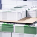 Mehrere exakt zugeschnittene Papierstapel liegen ordentlich auf einer Palette in einer Druckerei. Die Druckprodukte sind mit grünen Farbakzenten versehen und teilweise mit Papierbändern gebündelt. Auf der oberen Lage befindet sich ein zusätzlicher Stapel auf einer Zwischenlage aus Karton. Das Bild zeigt fertig produzierte Druckerzeugnisse der Druckerei Esslinger Druck in Villingen-Schwenningen, die für die Weiterverarbeitung oder Auslieferung vorbereitet sind.