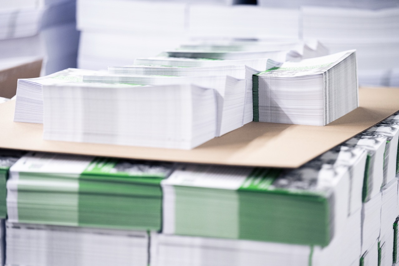 Mehrere exakt zugeschnittene Papierstapel liegen ordentlich auf einer Palette in einer Druckerei. Die Druckprodukte sind mit grünen Farbakzenten versehen und teilweise mit Papierbändern gebündelt. Auf der oberen Lage befindet sich ein zusätzlicher Stapel auf einer Zwischenlage aus Karton. Das Bild zeigt fertig produzierte Druckerzeugnisse der Druckerei Esslinger Druck in Villingen-Schwenningen, die für die Weiterverarbeitung oder Auslieferung vorbereitet sind.