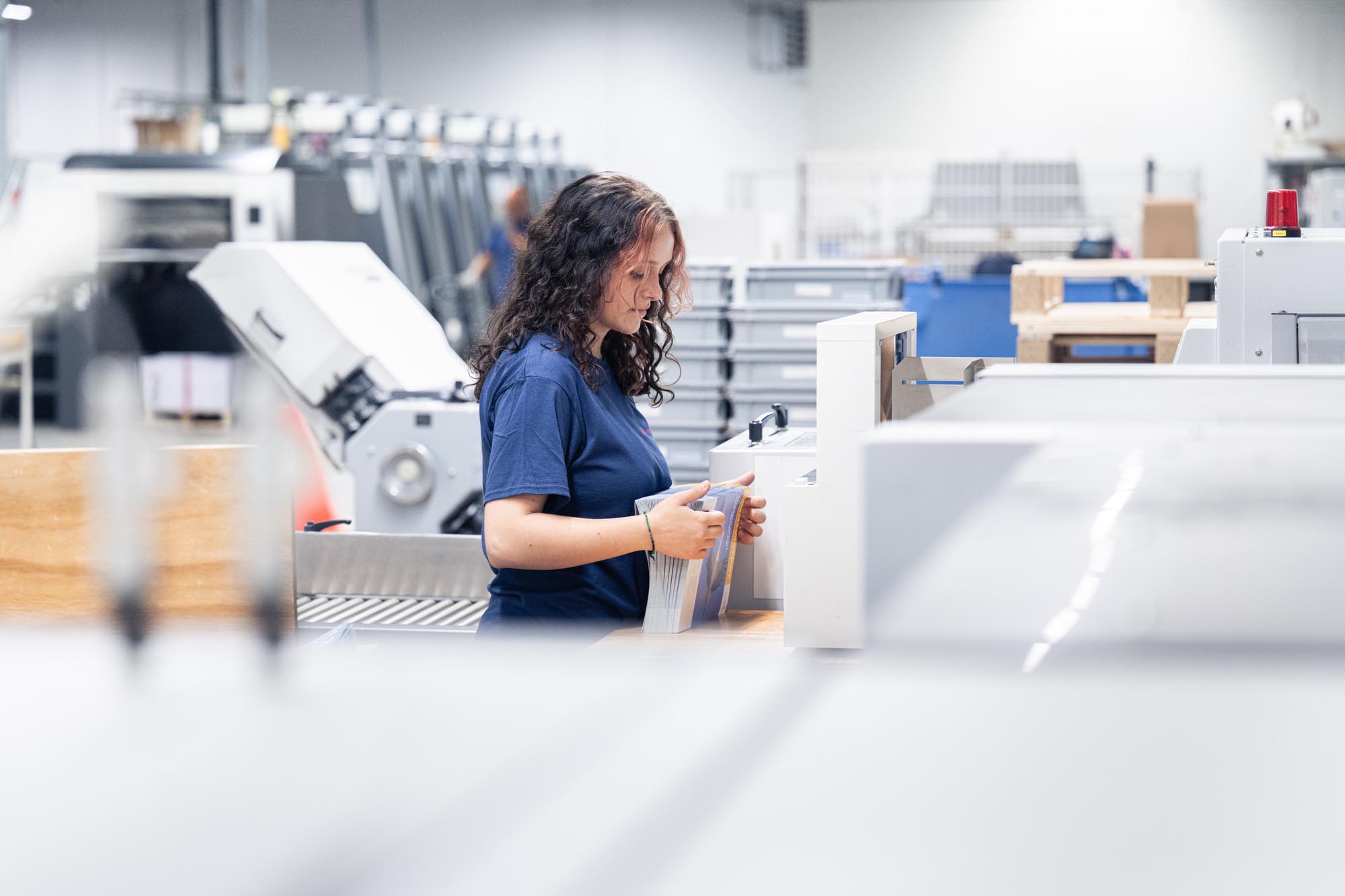 Eine Mitarbeiterin der Druckerei Esslinger Druck in Villingen-Schwenningen steht an einer industriellen Weiterverarbeitungsmaschine und hält einen Stapel frisch produzierter Mappen in den Händen. Sie trägt ein blaues T-Shirt und prüft konzentriert die ausgerichteten Druckprodukte vor der Weiterverarbeitung. Im Hintergrund sind weitere Druckmaschinen, Stapelboxen und Produktionsanlagen zu erkennen. Das Bild zeigt einen professionellen Arbeitsschritt in der Druckproduktion innerhalb einer hellen Fertigungshalle.