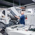 Ein Mitarbeiter der Druckerei Esslinger Druck in Villingen-Schwenningen öffnet die Abdeckung einer großformatigen Falzmaschine. Mit beiden Händen greift er in das geöffnete Maschinenmodul, um Einstellungen oder Wartungsarbeiten vorzunehmen. Die industrielle Anlage ist von Förderbändern, Metallkomponenten und transparenten Schutzabdeckungen umgeben. Das Bild zeigt einen technischen Arbeitsschritt in der professionellen Druckweiterverarbeitung.