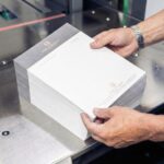 Hände eines Mannes halten einen Stapel Briefvorlagen mit grauem Kopfbereich und goldenem Logo auf einer Metallfläche einer Druckmaschine. Die Briefvorlagen wurden bei Esslinger Druck in Villingen-Schwenningen gedruckt.