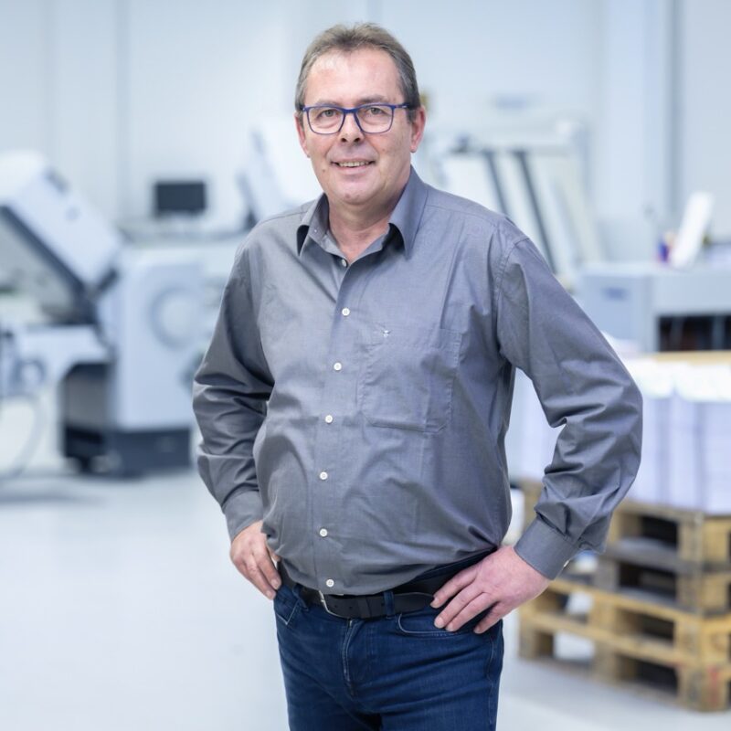 Ein lächelnder Mitarbeiter der Druckerei Esslinger Druck in Villingen-Schwenningen steht vor großen industriellen Druckmaschinen. Er trägt ein dunkelblaues Langarmshirt und graue Jeans und blickt selbstbewusst in die Kamera. Im Hintergrund sind Produktionsanlagen, Paletten und technische Geräte in einer hellen Halle zu sehen. Das Bild vermittelt Professionalität und Kompetenz im Bereich der Druckproduktion.