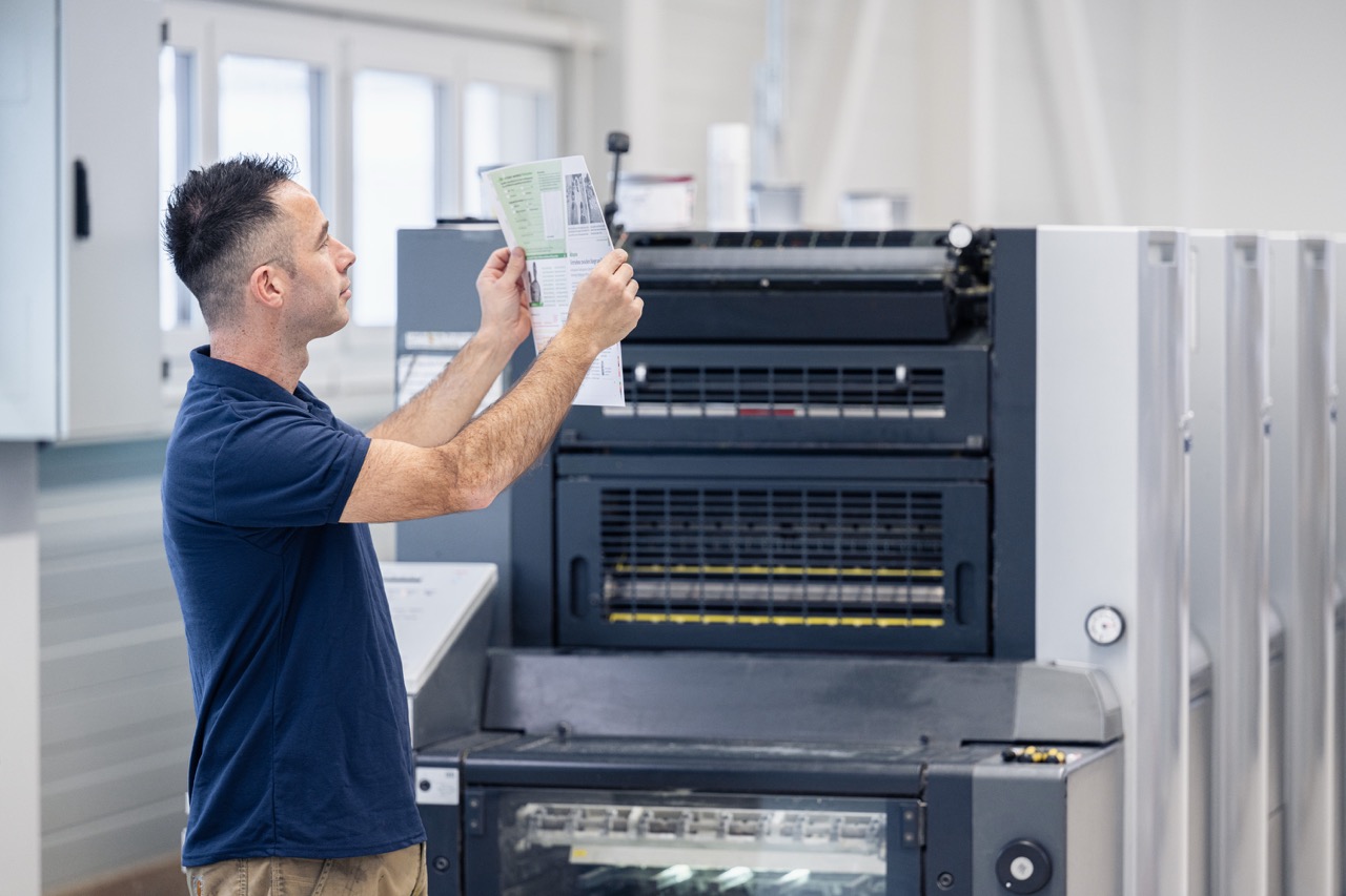 Ein Mitarbeiter der Druckerei Esslinger Druck in Villingen-Schwenningen steht vor einer großen Offsetdruckmaschine und hält einen frisch gedruckten Bogen prüfend gegen das Licht. Er kontrolliert aufmerksam Farbflächen, Textbereiche und Bilddetails, um die Druckqualität zu bewerten. Die industrielle Druckanlage mit mehreren Druckwerken befindet sich in einer hellen Produktionshalle. Das Bild zeigt die professionelle Qualitätskontrolle im Offsetdruck.