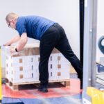 Ein Mitarbeiter der Druckerei Esslinger Druck in Villingen-Schwenningen beugt sich über eine Palette mit gebündelten Papierstapeln und fixiert diese mit transparenter Folie. Die Druckprodukte sind sauber gestapelt und mit Kartonzwischenlagen sowie Umreifungsbändern gesichert. Die Palette steht auf einem industriellen Boden innerhalb einer Produktionshalle mit Maschinen im Hintergrund. Das Bild zeigt die fachgerechte Verpackung von Druckerzeugnissen für Versand oder Lagerung.