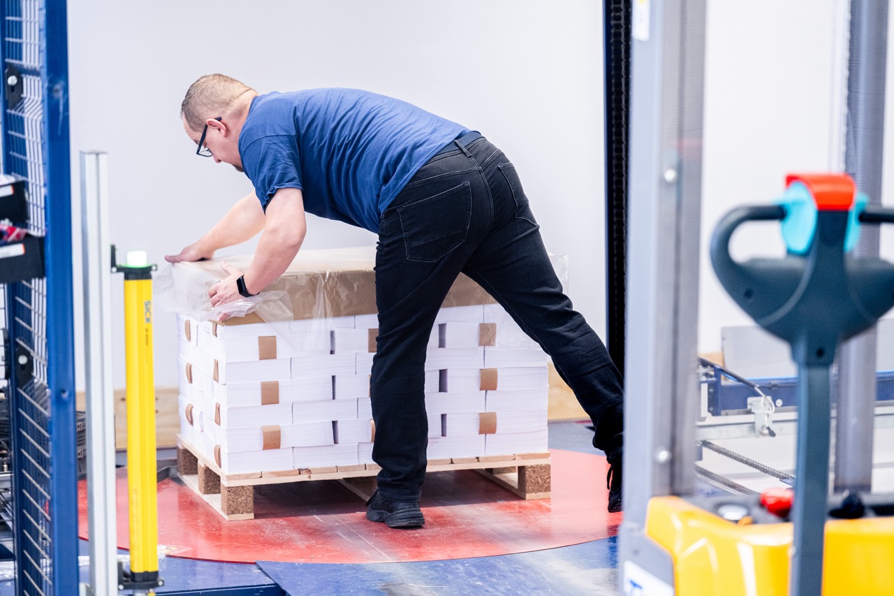 Ein Mitarbeiter der Druckerei Esslinger Druck in Villingen-Schwenningen beugt sich über eine Palette mit gebündelten Papierstapeln und fixiert diese mit transparenter Folie. Die Druckprodukte sind sauber gestapelt und mit Kartonzwischenlagen sowie Umreifungsbändern gesichert. Die Palette steht auf einem industriellen Boden innerhalb einer Produktionshalle mit Maschinen im Hintergrund. Das Bild zeigt die fachgerechte Verpackung von Druckerzeugnissen für Versand oder Lagerung.