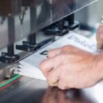 Detailaufnahme eines Mitarbeiters, der einen Stapel gefalzter Broschüren unter einer mehrspindeligen Lochmaschine exakt ausrichtet. Die Metallführungsschiene und die farbige Markierung auf dem Arbeitstisch sorgen für eine präzise Positionierung der Druckerzeugnisse. Oberhalb des Papiers sind die Bohrköpfe sichtbar, die für eine gleichmäßige Lochung vorbereitet sind. Die Szene veranschaulicht professionelle Papierverarbeitung in der modernen Druckerei Esslinger Druck in Villingen-Schwenningen.