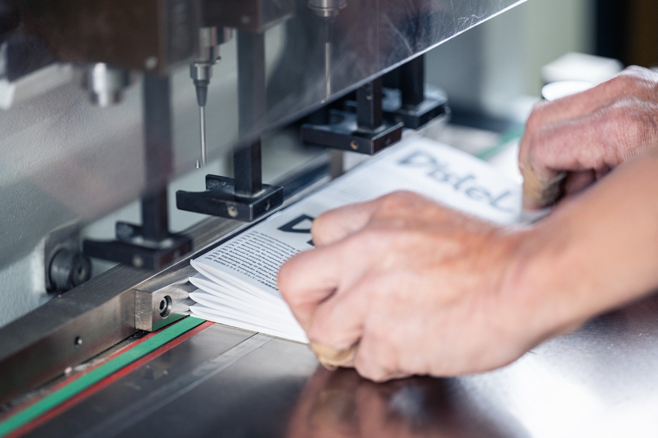 Detailaufnahme eines Mitarbeiters, der einen Stapel gefalzter Broschüren unter einer mehrspindeligen Lochmaschine exakt ausrichtet. Die Metallführungsschiene und die farbige Markierung auf dem Arbeitstisch sorgen für eine präzise Positionierung der Druckerzeugnisse. Oberhalb des Papiers sind die Bohrköpfe sichtbar, die für eine gleichmäßige Lochung vorbereitet sind. Die Szene veranschaulicht professionelle Papierverarbeitung in der modernen Druckerei Esslinger Druck in Villingen-Schwenningen.