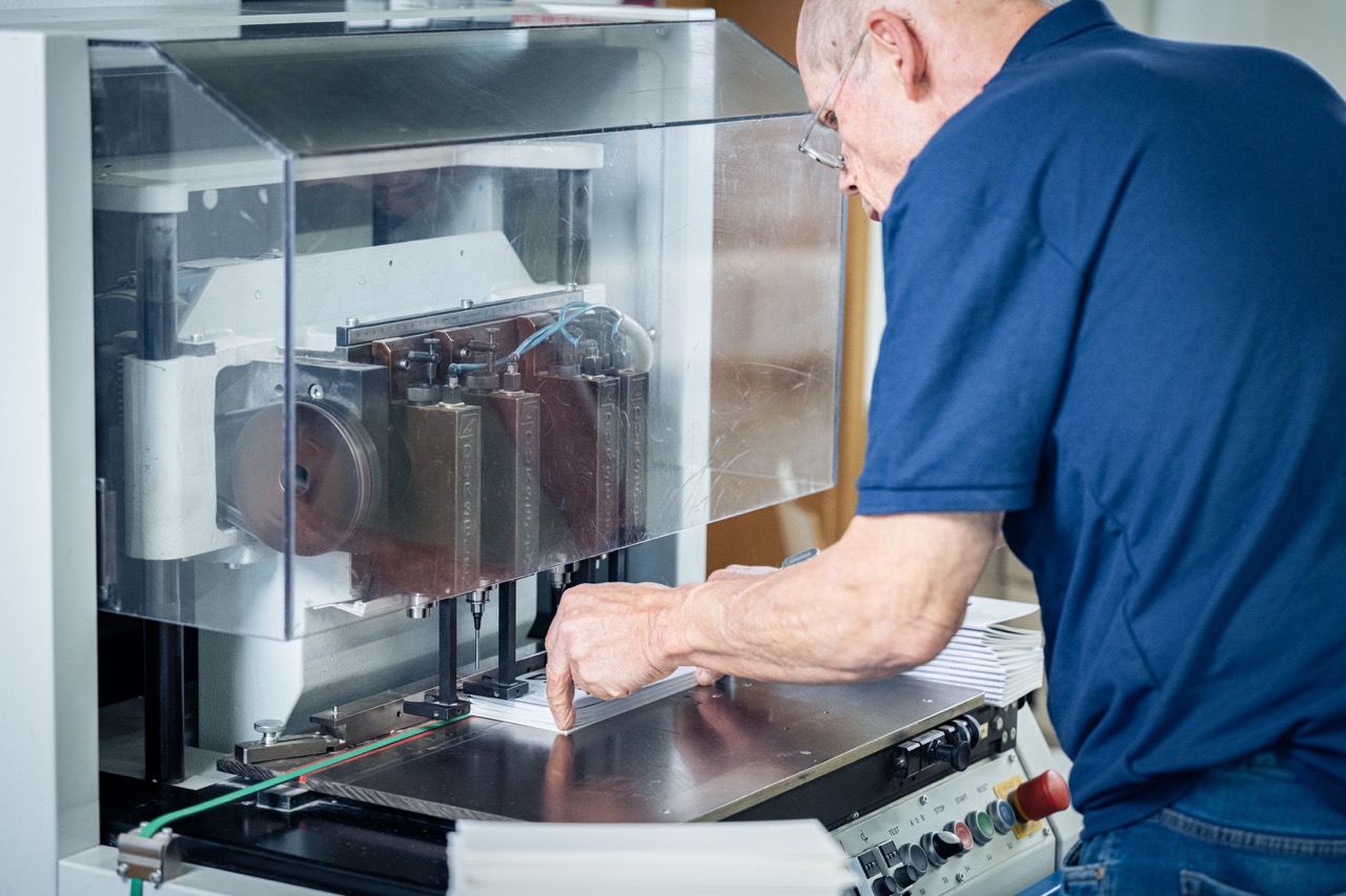 Ein älterer Mann mit Brille bedient eine industrielle Papierbohrmaschine in der Druckerei Esslinger Druck in Villingen-Schwenningen. Er richtet einen Stapel weißer Papierbögen präzise unter den Bohrköpfen aus, während die Maschine durch eine transparente Schutzabdeckung gesichert ist. Auf dem Arbeitstisch liegen weitere Papierstapel bereit, und Bedienelemente mit farbigen Knöpfen sind sichtbar. Die Szene zeigt einen professionellen Arbeitsschritt in der Weiterverarbeitung von Druckprodukten.