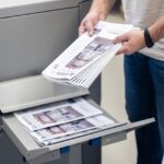 Eine Person entnimmt frisch gedruckte Werbeprospekte aus dem Ausgabeschacht einer großen Druckmaschine der Esslinger Druckerei in Villingen-Schwenningen. Die Hochglanz-Flyer mit Immobilienabbildung sind mehrfach gefaltet und liegen zusätzlich im Ablagefach der Maschine. Der Mann trägt ein schwarzes Armband und hält den Stapel fest in beiden Händen. Die Aufnahme zeigt einen typischen Arbeitsschritt beim professionellen Digitaldruck von Marketingmaterialien.