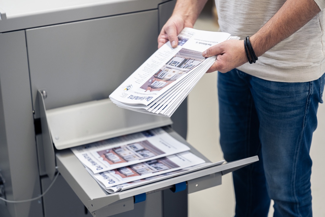 Eine Person entnimmt frisch gedruckte Werbeprospekte aus dem Ausgabeschacht einer großen Druckmaschine der Esslinger Druckerei in Villingen-Schwenningen. Die Hochglanz-Flyer mit Immobilienabbildung sind mehrfach gefaltet und liegen zusätzlich im Ablagefach der Maschine. Der Mann trägt ein schwarzes Armband und hält den Stapel fest in beiden Händen. Die Aufnahme zeigt einen typischen Arbeitsschritt beim professionellen Digitaldruck von Marketingmaterialien.