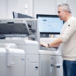 Ein Mitarbeiter steht in der Druckerei Esslinger Druck in Villingen-Schwenningen vor einer digitalen Produktionsdruckmaschine und bedient das integrierte Steuerpult. Auf dem Bildschirm hinter ihm sind Druckparameter und Produktionsdaten sichtbar, die zur Überwachung des Auftrags dienen. Die großformatige Maschine mit Papierzufuhr und Ausgabefach dominiert den Vordergrund der hellen Produktionsumgebung. Das Bild zeigt die professionelle Steuerung im Digitaldruck.