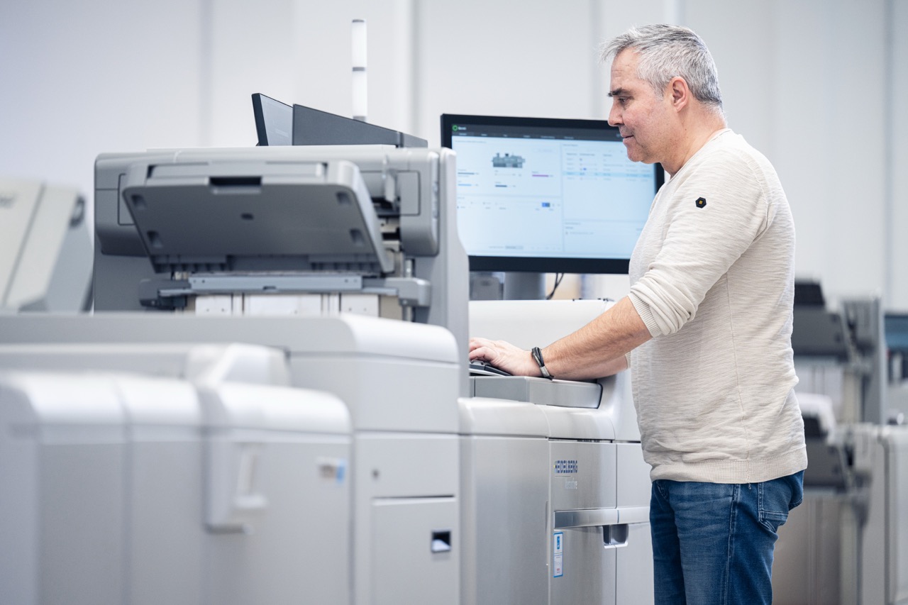 Ein Mitarbeiter steht in der Druckerei Esslinger Druck in Villingen-Schwenningen vor einer digitalen Produktionsdruckmaschine und bedient das integrierte Steuerpult. Auf dem Bildschirm hinter ihm sind Druckparameter und Produktionsdaten sichtbar, die zur Überwachung des Auftrags dienen. Die großformatige Maschine mit Papierzufuhr und Ausgabefach dominiert den Vordergrund der hellen Produktionsumgebung. Das Bild zeigt die professionelle Steuerung im Digitaldruck.