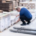 Ein Mitarbeiter kniet in einer Lagerhalle neben mehreren folierten Paletten mit gebündelten Druckprodukten und befestigt ein Versandetikett an einer Palette. Die Papierstapel sind mit Kunststofffolie umwickelt und mit grünen Umreifungsbändern gesichert. Im Hintergrund stehen weitere Paletten mit Kartonagen und verpackten Druckerzeugnissen ordentlich gestapelt. Das Bild zeigt die professionelle Versandvorbereitung im Bereich Logistik der Druckerei Esslinger Druck in Villingen-Schwenningen.