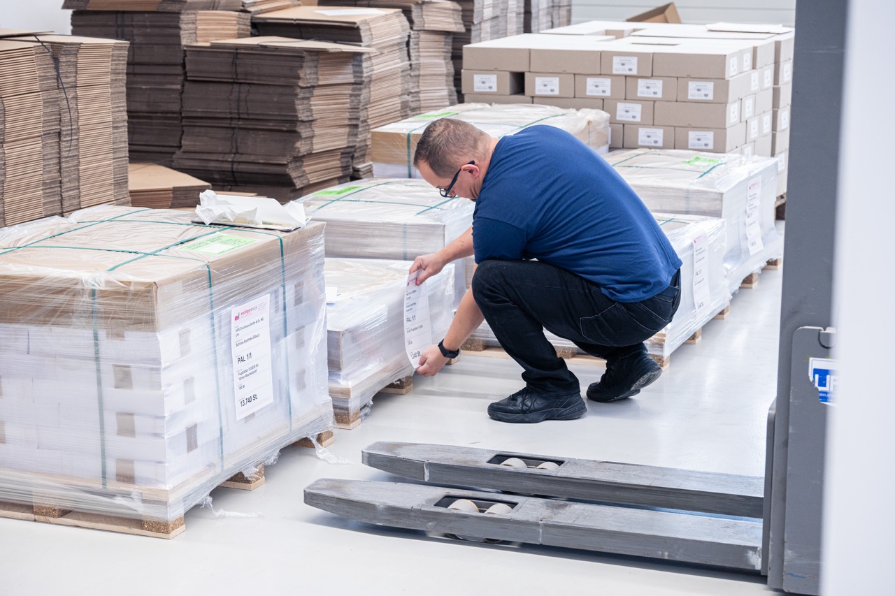 Ein Mitarbeiter kniet in einer Lagerhalle neben mehreren folierten Paletten mit gebündelten Druckprodukten und befestigt ein Versandetikett an einer Palette. Die Papierstapel sind mit Kunststofffolie umwickelt und mit grünen Umreifungsbändern gesichert. Im Hintergrund stehen weitere Paletten mit Kartonagen und verpackten Druckerzeugnissen ordentlich gestapelt. Das Bild zeigt die professionelle Versandvorbereitung im Bereich Logistik der Druckerei Esslinger Druck in Villingen-Schwenningen.