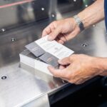 Hände halten zwei Visitenkarten über einem Stapel auf einer glänzenden Metalloberfläche. Die Visitenkarten sind weiß mit schwarzem Text und einem Logo auf einer Seite und dunkelgrau mit geprägtem Muster auf der anderen Seite. Die Visitenkarten wurden bei Esslinger Druck in Villingen-Schwenningen gedruckt.