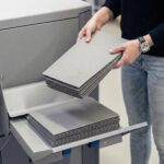 Eine Person hält einen Stapel grauer Mappen mit geprägtem Logo über dem Ausgabefach einer grauen Maschine, wo bereits weitere Dokumente liegen. Die Mappen wurden bei Esslinger Druck in Villingen-Schwenningen gedruckt.